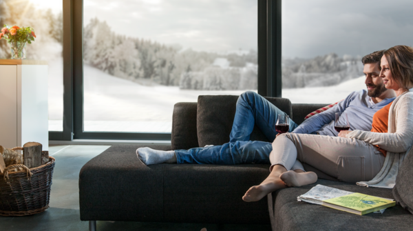 Personen sitzen entspannt auf einem modernen, dunklen Sofa vor einem knisternden Kaminfeuer, mit Blick auf eine verschneite Landschaft durch grosse Panoramafenster. Ein Korb mit Holz steht neben dem Kamin, Zeitschriften und Schuhe liegen im Wohnbereich.