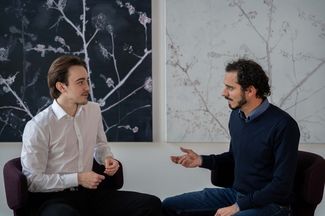 André Semadeni und Michael Naegeli sitzen während dem Interview vor zwei hängenden Bildern mit feinen Blumen.